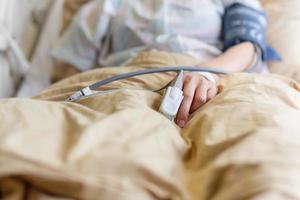 Asian patient's hand with an attached pulse oximeter on finger in hospital room. photo