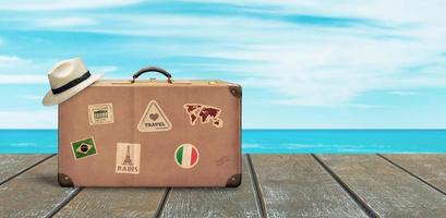 Old travel suitcase and white hat on wooden floor with sea and sky in background. Copy space beside. Tropical travel concept.