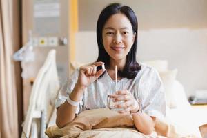 Woman holding white pills and a glass of water while sitting on a bed in hospital.