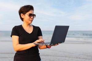 una mujer le disparó al cabello de pie en la playa cerca del océano sosteniendo una computadora portátil en la mano. foto