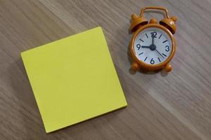 Yellow note with alarm clock on wooden desk. Flat lay photo