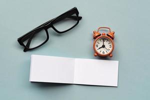 Notepad with reading glasses, pen and alarm clock on blue background. photo