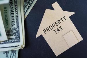 texto de impuestos sobre la propiedad en modelo de casa de papel marrón con billetes de dólar en un escritorio foto