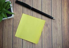 Yellow stick note on wooden table with potted plant and a pen. Directly above, flat lay for text. photo