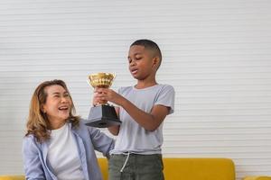Kid boy holding trophy and grandmother celebrating in living room