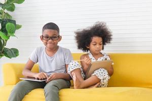 hermana y hermano jugando juntos en la sala de estar, dos niños pequeños hermanos sentados en un sofá sosteniendo una tableta y un libro