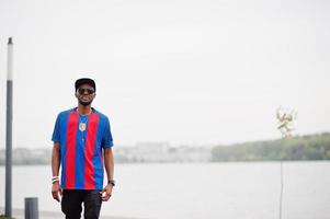 Stylish african american boy wear at cap, football t-shirt and sunglasses. Black sports man portrait. photo