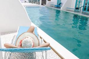 Back view of woman in summer hat laying on relaxing chair for sunbathing by the pool