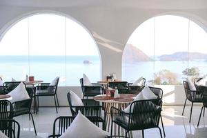 Tables and black chairs with pillow in dining area with sea view from window photo