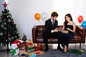 dulce pareja ama sonreír y pasar un tiempo romántico de navidad y celebrar la víspera de año nuevo en la decoración de un sofá marrón con árbol de navidad, globos coloridos y cajas de regalo en la sala de estar en casa foto