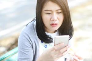 bella mujer asiática con vestido blanco se relaja con la mano sosteniendo un smartphone para socializar en línea en el parque. chica tailandesa y china. foto