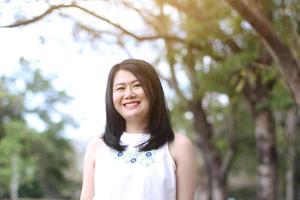 bella mujer asiática con vestido blanco se relaja y sonríe en el parque natural. chica tailandesa o chica china disfruta de vacaciones foto