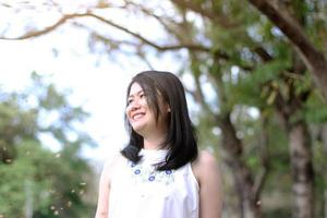 bella mujer asiática con vestido blanco se relaja y sonríe en el parque natural. chica tailandesa o chica china disfruta de vacaciones foto