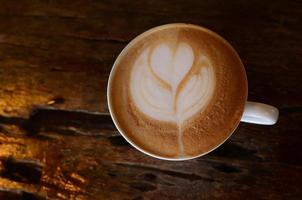 vista superior de una taza de café capuchino con leche caliente en una taza blanca sobre una mesa de madera, haga espuma de leche en forma de corazones y hojas. foto