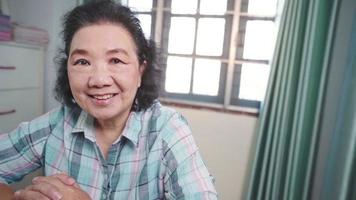 Asian senior woman facial expression sit down smiling looking at camera inside living room at working desk, showing happy confident face, healthy elderly having a wrinkle skin, an optimistic people video