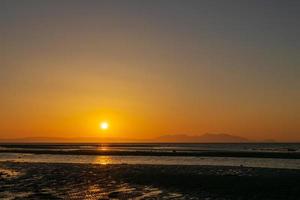 puesta de sol sobre la playa y la montaña, línea del horizonte reflejo de sombreado amarillo y naranja en el agua y la arena. enfocándose en la sombra de la superficie. paisaje naturaleza paisaje fondo. foto