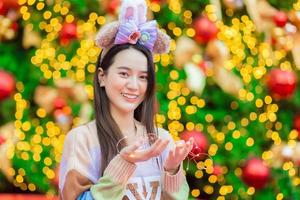 An Asian woman in a colorful sweater stands in front of the Christmas tree. With bokeh as background In the theme of celebrating the new year photo