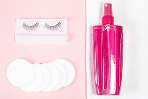 Makeup remover, white towel, cotton pads and false eyelashes on a pink background, top view