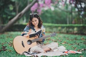 una joven asiática compone o escribe una canción y toca la guitarra en el parque, una mujer asiática escribe una canción con un cuaderno y toca la guitarra acústica sentada en el césped. foto