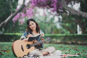 una joven asiática compone o escribe una canción y toca la guitarra en el parque, una mujer asiática escribe una canción con un cuaderno y toca la guitarra acústica sentada en el césped. foto