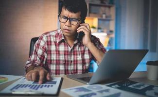 Freelance asian man with stressed and worried expression Talking on mobile phone and working on a laptop use hand to point the graph. freelancers are worried unfinished work, thinking photo