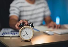 Selective focus of alarm clock and freelance man pressing alarm clock button while working at home. Business man working overtime at night in the home. photo