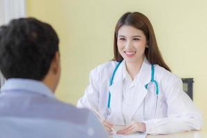 una hermosa doctora asiática sonrió y preguntó acerca de los síntomas. quedarse en el hospital con una bata blanca y un estetoscopio foto
