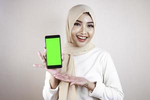 Portrait of cheerful young Muslim Asian woman pointing to cellphone with green screen photo