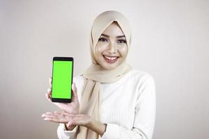 Portrait of cheerful young Muslim Asian woman pointing to cellphone with green screen photo