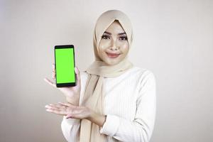 Portrait of cheerful young Muslim Asian woman pointing to cellphone with green screen photo