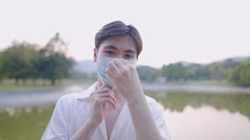 Close up portrait gesturing of asian young man in white shirt put on and unfold mask with thumbs up isolated both hands, while standing next to pond park in city Confidence boosting after epidemic video