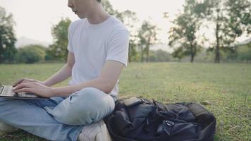 ein attraktiver asiatischer mann, der auf einer wiese sitzt und tagsüber mit einem laptop im grünen park arbeitet, fernarbeit an einem inspirierenden grünen ort, ein multitasking-arbeiter, der tippt und telefonanrufe entgegennimmt video