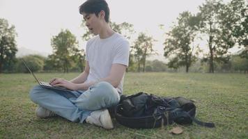 Homem asiático de pele clara bonito trabalhando com laptop, sente-se na grama, local de trabalho remoto do parque ao ar livre, luz diurna de bom tempo, tecnologia digital, trabalhador freelance criativo, roupas casuais