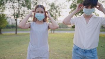 Asian male and female put on medical face mask looking straight at camera, protection from covid pandemic illness, infectious diseases prevention, shot during sunset golden hour self awareness caution
