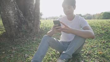 jeune bel homme assis tenant un smartphone, se détendre sous l'ombre des arbres à l'intérieur du parc, journée chaude et ensoleillée, style de vie moderne, technologie numérique, gadget portable, travail à distance en plein air