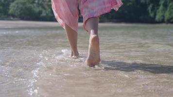 pernas e pés femininos caminhando ao longo da praia da ilha com salpicos de ondas de água salpicos de água e com os pés descalços em câmera lenta. vista por trás de baixo ângulo da câmera, seguindo a mulher andando na praia