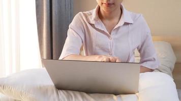 arbetande kvinna som arbetar med bärbar dator och letar efter forskningsmaterial online, sitter på sängen med en bekväm kudde i knät, varmt morgonsolljus läcker genom tunna vita bomullsgardiner video