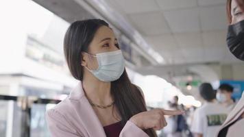 Close up Asian office working women wear medical face mask standing on platform and having talk while waiting for their train arriving, infectious coronavirus prevention, respiratory system protection