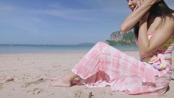 Young attractive asian black hair girl talking on phone while sitting on a clean beachfront, summer vacation, tropical zone travelling, using phone to communicate, shy reaction when falling in love video