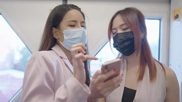 Two asian female closed colleague standing cheerfully talking while using a pubic train to office, attractive red dyed hair fashionista shows smartphone to friend, people working on covid-19 pandemic video