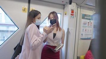 Two females colleagues friends wear protective face mask standing inside metro sky train, girls going to work together, public transportation, discussing on the work project, women in business attire video