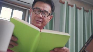 Senior asian man wear eye glasses reads book tapping on tablet at home living room desk, focusing concentrate while reading, old age learning, reading stories relaxation, middle age free time hobbies video