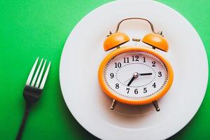 Orange vintage style alarm clock on a white plate with a fork. Simple meal time concept flat lay.