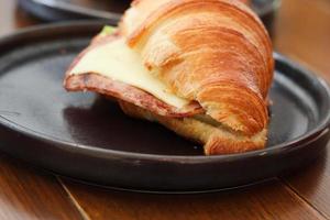 Delicioso sándwich de croissant casero recién horneado con queso de jamón en un plato negro sobre una mesa de desayuno de madera. foto