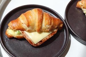 Delicioso croissant casero recién horneado con queso de jamón en un plato negro sobre una mesa de desayuno de madera, vista superior de la mesa de desayuno. foto