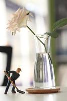 White rose in a glass vase with miniature worker doll holding shovel on table, miner man at work tiny figure toy model digging ground or gardening photo