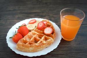 gofres vieneses con fresa y plátano en plato blanco con jugo de naranja en mesa oscura. foto