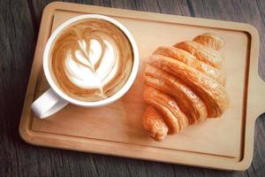 Delicioso croissant recién horneado con una taza de hermoso café matutino en una tabla de cortar de madera, vista superior de la mesa del desayuno.