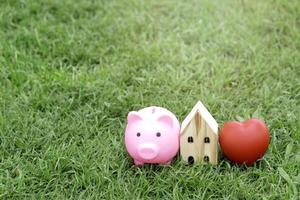 alcancía rosa, corazón rojo y casa de madera sobre hierba verde, ahorra dinero para una casa foto