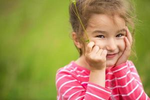 una niña pequeña con una camiseta a rayas de coral sobre un fondo verde en un campo sostiene su cara en sus manos y sonríe astutamente. día del niño, niño feliz, protección del medio ambiente y la naturaleza, repelente de insectos